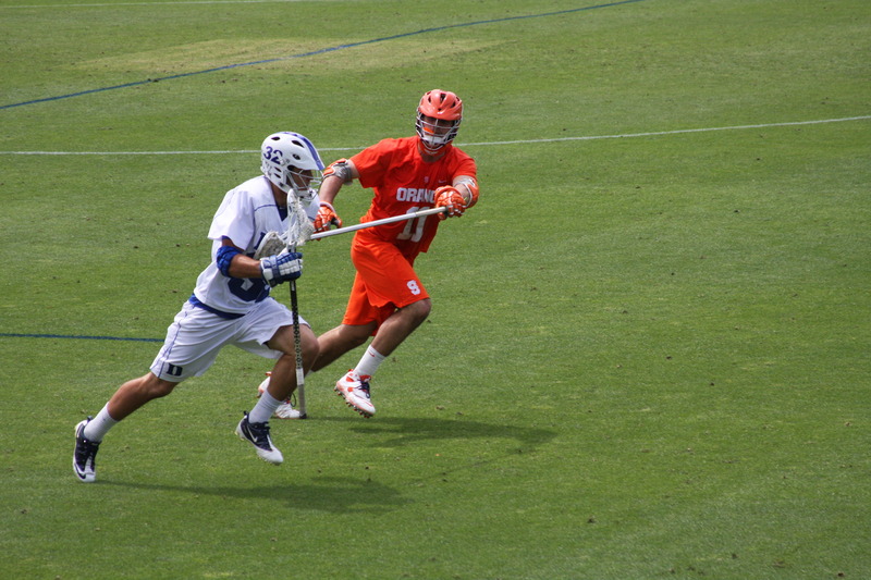 On an athletic field a Syracuse University defender prods a Duke University midfielder with his lacrosse stick